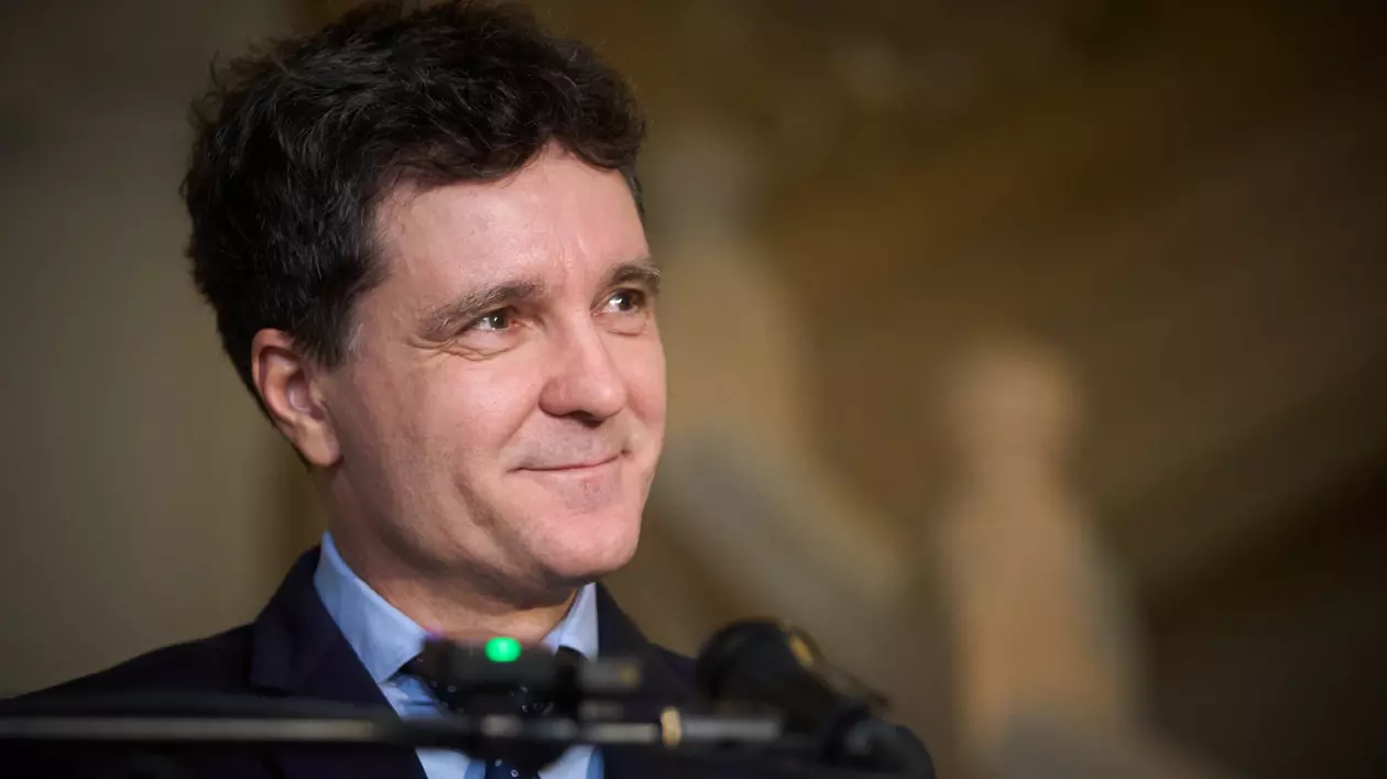 Nicusor Dan, la o conferință de presă, cu un microfon pus pe un stativ in fata sa. E imbracat intr-un sacou bleumarin, cămasă bleu și cravată albastră.